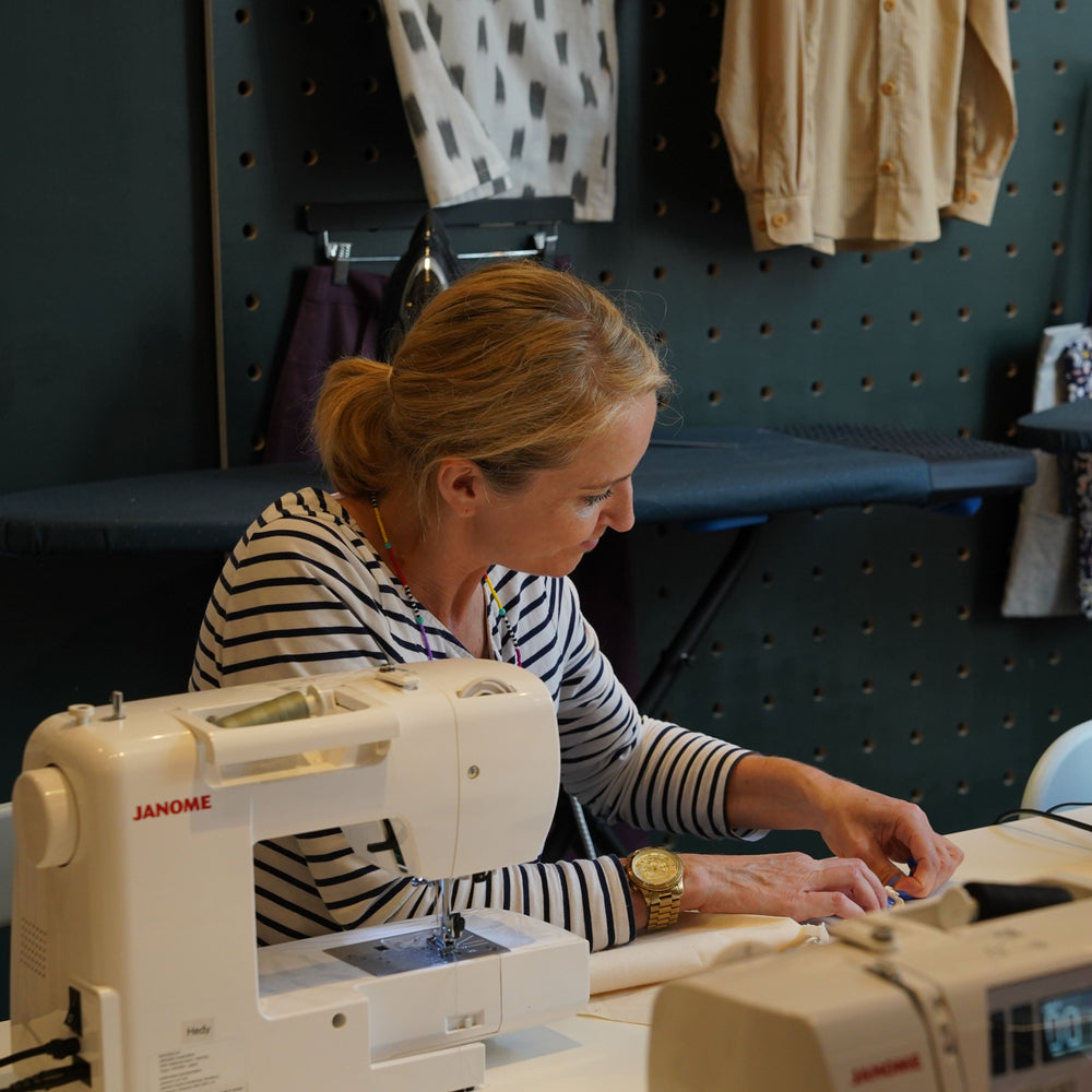 Woman working on a sewing machine in a workshop setting with various bags and materials in the background.