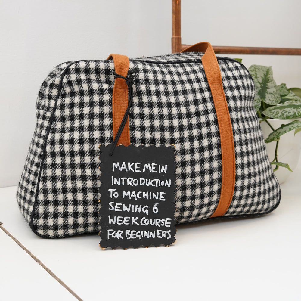 Plaid holdall with brown straps on a white surface, accompanied by a sign about a sewing course.